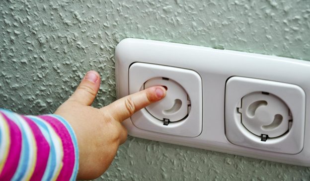 La main de l'enfant dans une manche rayée touche une prise de courant sur un mur texturé ; priorité à la sécurité des enfants.