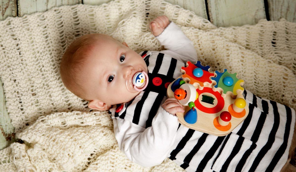 Bébé, 8 à 12 semaines, en tenue rayée, tétine bien ajustée, tient un jouet coloré sur une couverture tricotée.