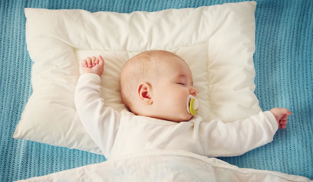 Un bébé dort paisiblement sur un lit superposé, portant une tétine sous une couverture légère sur une surface texturée bleue.